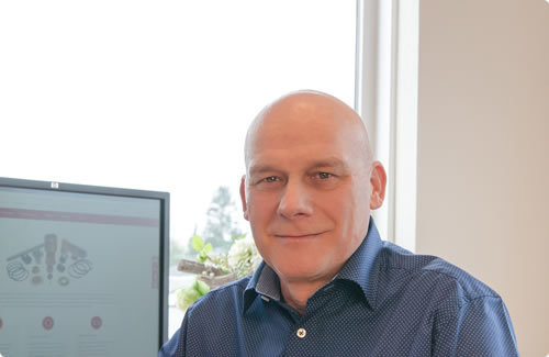 Freundlich blickender Mann mit Glatze am Schreibtisch, im Hintergrund ein Monitor mit technischer Website und ein Fenster mit Tageslicht